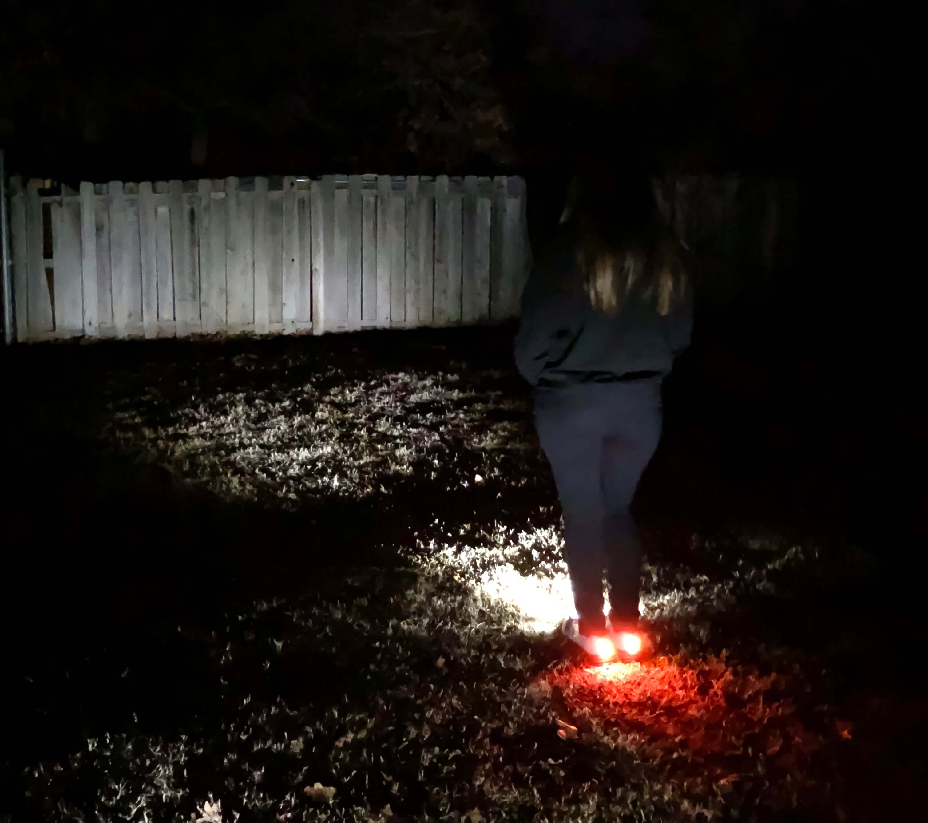 A woman wearing illuminated night runner shoes in a dark yard, highlighting safety lights for evening visibility.
