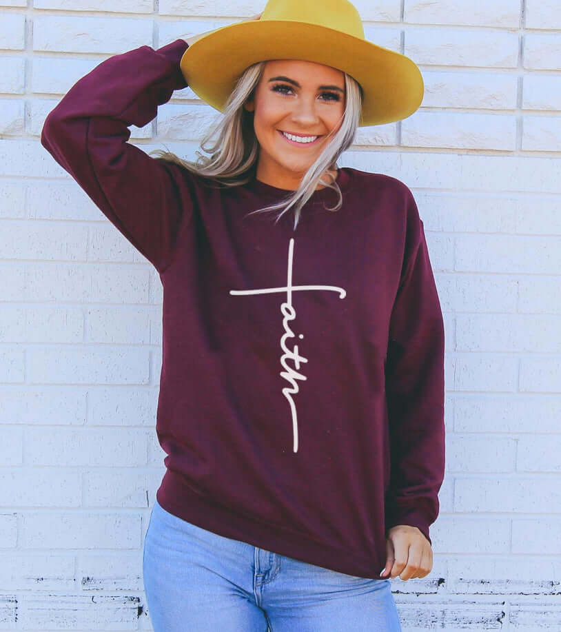 Stylish woman wearing a maroon Faith Sweatshirt with a white graphic, accessorized with a yellow hat.